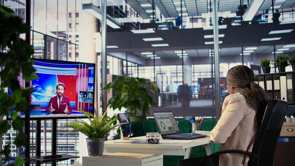 Woman in an office watching a news broadcast on a screen